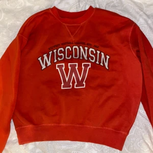 Röd Wisconsin college-tröja - Snygg röd sweatshirt med stort Wisconsin-tryck och en stor vit W på bröstet. Klassisk college-stil med rund hals och ribbade muddar. Perfekt för dig som gillar sportig och avslappnad look.