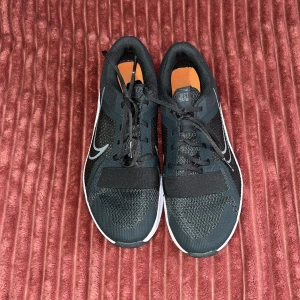 Svarta Nike sneakers med meshdetaljer - Snygga svarta Nike sneakers med vit sula och grå swoosh på sidan. Ovandelen är i mesh och syntet för extra andningsförmåga. Skorna har snörning och en sportig vibe, perfekta för träning eller chill vardagsstil.