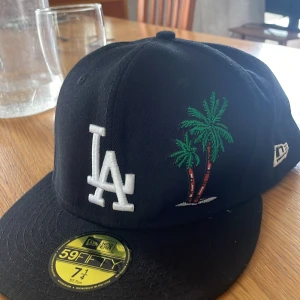 LA Dodgers svart keps New Era 59FIFTY - Svart LA Dodgers keps från New Era 59FIFTY med broderad vit LA-logga framtill och palmer i grönt och brunt. På sidan finns ett 1988 World Series-märke. Klassisk flat peak och broderade detaljer. Perfekt för dig som gillar sportig streetstyle.