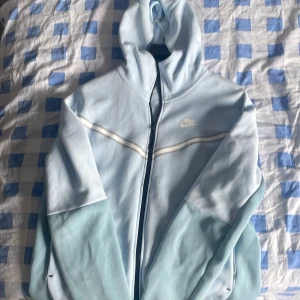 Ljusblå Nike hoodiejacka med dragkedja - Nike hoodiejacka i ljusblått med hel dragkedja och huva. Jackan har vita detaljer över bröstet och är tillverkad i mjukt material, perfekt för chill eller träning. Snygg och enkel design med diskret Nike-logga på bröstet.
