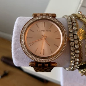 Guldig klocka från Michael Kors - Snygg guldig klocka från Michael Kors med rund urtavla och glittrande stenar runt boetten. Klockan har en länkad metallrem och stilren design med minimalistiska markeringar på urtavlan. Perfekt accessoar för att lyfta din stil.