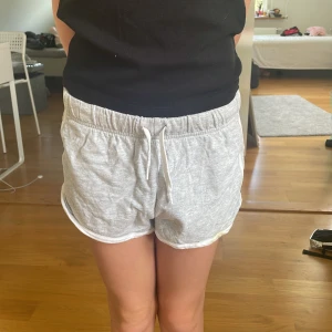 Grå mjuka shorts från H&M  - Superbekväma ljusgrå shorts från H&M med vit kant och snörning i midjan i storlek 134. Perfekta för sommardagar eller chilla hemma. Tillverkade i mjuk bomull och har en loose fit som gör dem extra sköna att bära. Har en gul fläck på framsidan men syns inte så mycket. 