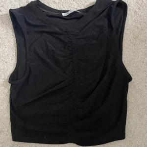 Svart croppad linnetopp från Zara - Svart croppad linnetopp från Zara med snygg rynkdetalj framtill. Toppen är ärmlös och har en enkel, stilren design som passar perfekt till jeans eller kjol. Mjuk och stretchig för en bekväm känsla.