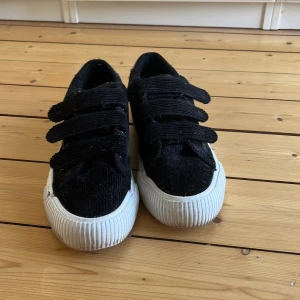 Svarta manchestersneakers Even and odd skor - Snygga svarta sneakers i manchestertyg med breda kardborreband och vit, räfflad gummisula. Skorna har rund tå och en blank detalj på hälen som ger extra edge. Perfekta för dig som gillar en stilren men cool look.Har använts 2 gånger och är i nytt skick iprincip 