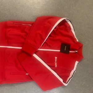 Röd Palm Angels vindjacka med vita detaljer - Riktigt snygg röd vindjacka från Palm Angels med vita ränder längs ärmarna och dragkedja framtill. Jackan har hög krage, två fickor med dragkedja och logga broderad på bröstet. Perfekt för dig som vill sticka ut med streetwear-vibe.