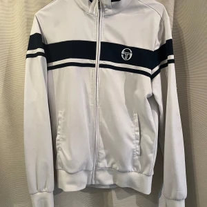 Vit fleece jacka från Sergio Tacchini - Vit fleece jacka från Sergio Tacchini med blåa ränder över bröstet och ärmarna. Jackan har dragkedja framtill, ståkrage och två fickor. Ikonisk logga broderad på bröstet och detaljerad zip puller. Perfekt för en sportig och retro vibe.