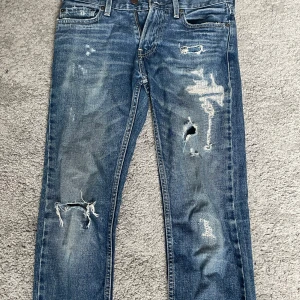 Hollister skinny jeans med slitningar - Snygga blå skinny jeans från Hollister med slitna detaljer och hål på benen för en cool, avslappnad look. Klassisk femficksmodell i stretchig bomullsdenim. Perfekta för dig som gillar streetstyle och vill ha ett par jeans med attityd.