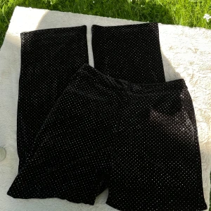 Svarta prickiga sammetsbyxor - Säljer ett par svarta byxor i mjuk sammet med små silvriga stenar över hela tyget. Byxorna har raka, vida ben och stängs med dragkedja och hake i midjan. Perfekta för dig som vill sticka ut lite extra med ett glittrigt mönster.
