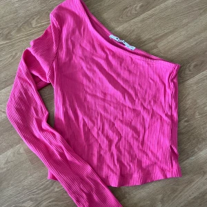 Ribbad rosa oneshoulder topp - Säljer en ribbad rosa topp från NA-KD med oneshoulder-design och lång ärm. Toppens material är stretchigt och sitter snyggt på kroppen. Perfekt för dig som vill sticka ut med en färgstark och trendig look.