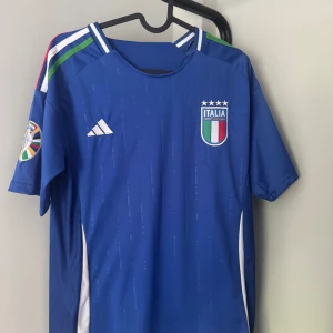 Italien Adidas fotbollströja blå - Snygg blå fotbollströja från Adidas med Italiens landslagsmärke på bröstet och vita detaljer längs sidorna. Tröjan har korta ärmar, klassiska Adidas-ränder på axlarna i grönt, vitt och rött samt broderad logga. Perfekt för fotbollsfans!