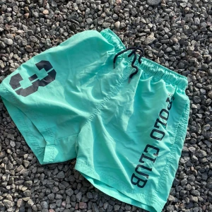 Mintgröna badshorts Vinson Polo Club - Fräscha mintgröna badshorts från Vinson Polo Club med svart snörning i midjan, stort tryck med siffran 3 på ena benet och POLO CLUB-logga längs sidan. Innerbyxa i mesh för extra komfort. Perfekta för stranden eller poolen!