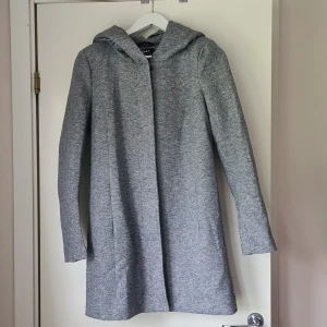 Grå kappa med huva från ONLY - Snygg grå kappa från ONLY med huva och stilren design. Jackan har dragkedja och knappar framtill, lång ärm och en rak passform. Perfekt för dig som gillar en enkel men trendig look, passar bäst under höst och vår då den inte är fodrad. Jackan är oanvänd då jag tyckte att den var för trång över armarna, har dock lyckats dra loss lappen. Supersnygg, så jag är lite besviken över att den aldrig kom till användning.. 