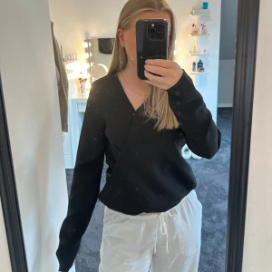 Svart omlottblus med lång ärm - Säljer en svart omlottblus med lång ärm och v-ringning. Blusen har en stilren look med knäppning på insidan för att hålla omlottstilen på plats. Perfekt för dig som gillar minimalistiska och trendiga plagg.