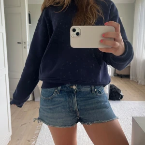 Blå jeansshorts med fransig kant - Snygga blå jeansshorts med fransig, rå kant och klassisk femficksdesign. Shortsen har normal passform och bälteshällor, perfekta för en chill sommarlook. De är tillverkade i denim och har en avslappnad vibe med slitna detaljer längst ner. Midjemått: 76 cm