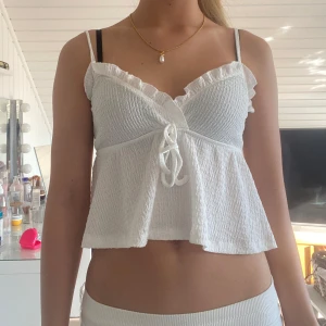 Vit volangtop med knyt och smock - Supersöt vit linnetopp med smockad överdel, volangkant vid bysten och tunna axelband. Toppen har omlottkänsla framtill och knyt i mitten. Luftig och flowy passform, perfekt för varma dagar.