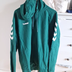 Grön Hummel hoodie med tryck XL - Snygg grön hoodie från Hummel i storlek XL. Tröjan har vit Hummel-logga på bröstet och vita pilar längs ärmarna. Utrustad med huva och dragsko, samt tillverkad i mjuk bomull. Perfekt för en sportig och avslappnad stil.