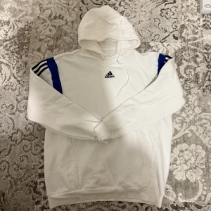 Vit Adidas hoodie med blå detaljer - Vit hoodie från Adidas med svarta och blå ränder på axlarna och broderad logga på bröstet. Tröjan har huva med snörning och är långärmad. Perfekt för dig som gillar sportig och clean stil.