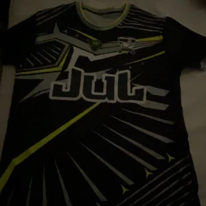 Svart Yamaha t-shirt med grafiskt tryck - Cool svart t-shirt med stora grafiska tryck i vitt och neongult, både fram och bak. Framsidan har ett futuristiskt mönster och texten 'Jul', medan baksidan har handmotiv och Yamaha-logga. Perfekt för dig som gillar streetwear och motorsport.