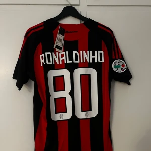 AC Milan Ronaldinho tröja Adidas - Säljer en klassisk AC Milan fotbollströja med Ronaldinho och nummer 80 på ryggen. Tröjan är svart och röd med vertikala ränder, Adidas-logga på bröstet och Bwin som sponsor. Serie A-märke på ärmen och klubbmärke framtill. Perfekt för dig som älskar fotboll och retrostil.