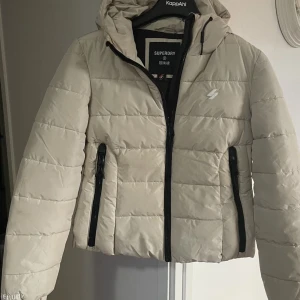 Beige pufferjacka från Superdry - Snygg beige pufferjacka från Superdry med huva och svarta dragkedjor. Jackan är quiltad och har två fickor framtill med dragkedja. Perfekt för kyliga dagar och riktigt skön att bära. Klassisk Superdry-logga på bröstet.