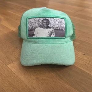 Velvet keps med Péle tryck från TIARIS - Snygg mintgrön keps från TIARIS med mesh på sidorna och baksidan. Framtill finns ett svartvitt retro fototryck av en fotbollsspelare. Kepsen har böjd skärm och justerbar passform. Perfekt för dig som vill sticka ut med en unik accessoar.