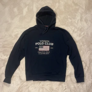 Mörkblå hoodie från Vinson Polo Club - Mörkblå hoodie från Vinson Polo Club med vit text och amerikansk flagga framtill. Tröjan har huva med snörning och långa ärmar. Perfekt för dig som gillar klassisk och sportig stil.