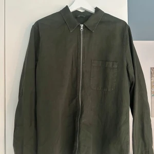 Olivgrön overshirt från A Day's March - Säljer en stilren olivgrön overshirt från A Day's March. Jackan har klassisk krage, bröstficka och hel dragkedja framtill. Perfekt lager-på-lager-plagg med enkel, clean look och lång ärm. Ordinariepris 1400 kr.