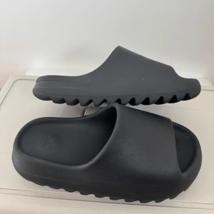 Svarta adidas Yeezy Slides - Svarta adidas Yeezy Slides med chunky sula och minimalistisk design. Tillverkade i syntetmaterial för en mjuk och bekväm känsla. Perfekta för chill dagar och har den ikoniska Yeezy-loggan på insidan samt adidas-logga i sulan.