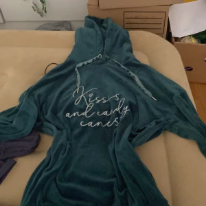 Mörkgrön velourhoodie Hunkemöller S - Mysig mörkgrön hoodie från Hunkemöller i mjuk velour med huva och snörning. På bröstet står det 'Kisses and candy canes' broderat i vitt. Perfekt för chill dagar när du vill vara extra comfy.