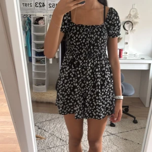 Svart blommig kortklänning med inbyggda skorts - Supersöt svart kortklänning med inbyggda shorts, med vita små blommor, puffärmar och fyrkantig ringning. Klänningen har en flowy passform och är perfekt för varma dagar. Materialet känns lätt och luftigt, och den har en smickrande midja. 