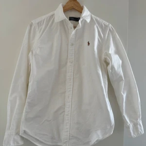 Vit skjorta från Polo Ralph Lauren - Klassisk vit skjorta från Polo Ralph Lauren i 100% bomull. Skjortan har lång ärm, knappar framtill och den ikoniska broderade polospelaren på bröstet. Perfekt för en clean och stilren look.