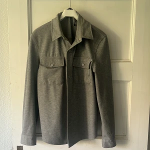 Grå overshirt med knappar - Stilren grå overshirt med klassisk krage och tryckknappar framtill. Två bröstfickor med lock och knappar ger en snygg och praktisk look. Långärmad modell i mjukt material, perfekt att slänga över en t-shirt för en chill vibe.