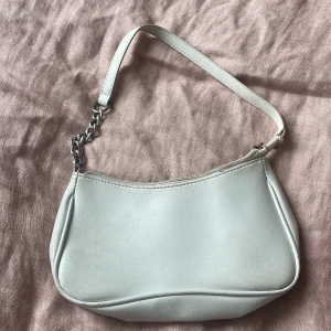 Vit axelväska med kedjedetalj - Säljer en stilren vit axelväska i syntetmaterial med silverfärgad kedjedetalj på axelremmen. Väskan har dragkedja upptill och är ca 25 cm bred och 14cm hög. Enkel och clean design som passar till många outfits.