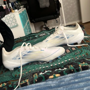 Vita Adidas fotbollsskor med blå detaljer - Säljer ett par snygga vita Adidas fotbollsskor med blå och ljusgula detaljer. Skorna har snörning och är tillverkade i syntetmaterial för lätt känsla på planen. Perfekta för dig som vill sticka ut med stilrena och moderna skor på matchen. Säljs för att dem va för små på mig trodde dem skulle passa storlek på dem är 43 1/3. Köptes för 1699 kr från unisport 