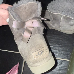 Grå UGG boots med rosett - Mysiga grå UGG boots i mocka med fluffigt foder och dekorativa rosetter baktill. Skorna har rund tå och platt sula, perfekta för kalla dagar. Klassisk UGG-logga på hälen och en stilren look som passar till många outfits. Köpt för ca 2500, däremot mycket välanvända. Går att tvätta och skrubba vilket gör att de kmr se fräschare ut men det är eget arbete.