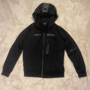 Svart tracksuit tröja från Hoodrich - Svart tracksuit tröja från Hoodrich med tryck på bröstet, ryggen och huvan. Jackan har hel dragkedja, huva och en bröstficka med dragkedja. Tillverkad i slät polyester med sportig vibe och ribbade muddar. Perfekt för dig som gillar streetwear och snygga detaljer.