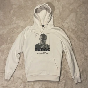 Vit Tupac hoodie från Mister Tee - Vit hoodie från Mister Tee med stort Tupac-tryck och texten 'ONLY GOD CAN JUDGE ME' på bröstet. Klassisk känguruficka framtill, huva med dragsko och långa ärmar. Perfekt för dig som gillar streetwear och vill sticka ut med en ikonisk look. Enda negativa är att de finns sminkfläck vid luvan