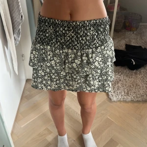 Blommig volangkjol från Zara - Supersöt kort kjol från Zara med svart och vit blommig print. Kjolen har smock i midjan och volangdetalj som ger en flowy känsla. Perfekt för dig som gillar en feminin och trendig stil. Storleken är 152 men sitter som en xs