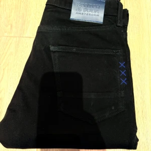 Svarta jeans Scotch & Soda Ralston - Svarta jeans från Scotch & Soda, modell Ralston. Snygga blå detaljer med tre korsstygn vid bakfickan och blå sömmar på insidan. Klassisk femficksmodell i stretchig bomullsblandning för skön passform.