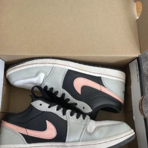 Nike Air Jordan 1 Low grå/svart/rosa - Nike Air Jordan 1 Low sneakers med svart och grå ovandel i skinn, rosa swoosh och detaljer. Klassisk Air Jordan-logga på hälen och svart snörning. Perforerad tåbox för extra ventilation. Snygg och stilren färgkombo som sticker ut.