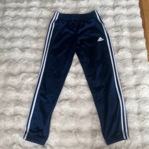 Adidas marinblå träningsbyxor - Marinblå träningsbyxor från Adidas med klassiska vita ränder längs sidorna och Adidas-logga på låret. Byxorna har resår i midjan med blå dragsko och är gjorda i glansig polyester. Perfekta för sport eller chill.