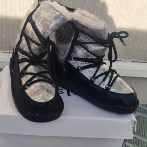 Silvriga moon boots med svart mocka strl 37 - Snygga moon boots i metallic silver med svarta mockadetaljer och svarta snören. Insidan är fodrad med mjuk päls för extra värme. Skorna har en cool, glittrig look och passar perfekt för vinterns kalla dagar.