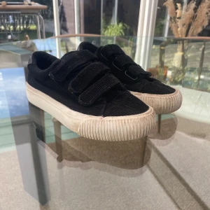 Svarta manchestersneakers med kardborre - Svarta sneakers i manchestertyg med tre breda kardborreband och vit, räfflad gummisula. Skorna har rund tå och en cool detalj i blankt material på hälen. Perfekta för en avslappnad och trendig look.