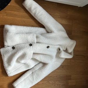 Vit teddyjacka från Zara Kids - Supermysig vit teddyjacka från Zara Kids med stora bruna knappar framtill. Jackan har lång ärm, hög krage och är gjord i fluffigt teddy-material som håller dig varm under kyliga dagar. Perfekt för dig som vill ha en stilren och trendig look. Den passar även M