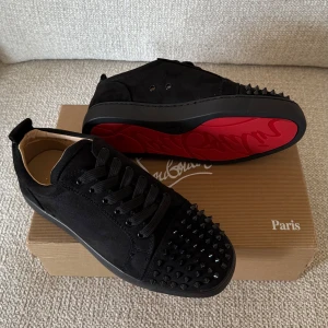 Christian Louboutin svarta sneakers med nitar - Svarta sneakers i mocka från Christian Louboutin med ikoniska röda sulor och svarta nitar på tån. Skorna har snörning och en stilren, rund tå. Perfekt för dig som vill sticka ut med en edgy och lyxig vibe.