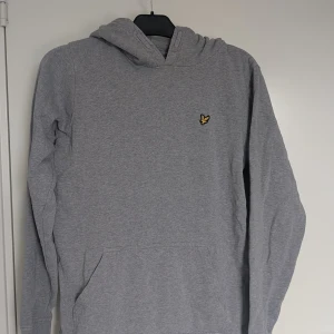 Grå hoodie från Lyle & Scott Junior ca 170 - Snygg grå hoodie från Lyle & Scott Junior med klassisk gul logga på bröstet. Tröjan har huva, känguruficka och är tillverkad i mjuk bomullsmix. Storleken är 15-16 år. Motsvarar storlek 170 eller XS/S i herr