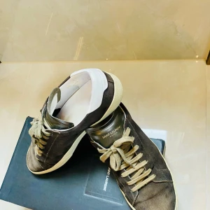 Bruna Saint Laurent sneakers i mocka - Snygga bruna sneakers från Saint Laurent i mjuk mocka med beige snörning och vit häl. Klassisk låg modell med rund tå och platt sula. Perfekta för dig som gillar stilrena och exklusiva skor med en lyxig känsla.