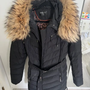 Svart lång dunjacka Hollies  - Säljer en svart lång dunjacka från Hollies med stor huva och bred päls i beige/bruna toner. Jackan har dragkedjor, flera fickor och markerad midja med bälte. Perfekt för kalla dagar och riktigt snygg med sitt quiltade mönster och detaljer.  Äkta pälskrage.