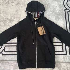 Svart Burberry hoodie  - Svart hoodie från Burberry med klassiskt rutigt foder i huvan och snygga snörningsdetaljer. Tröjan har dragkedja framtill, två fickor och diskret Burberry-logga på bröstet. Perfekt för dig som vill ha en stilren och lyxig look.