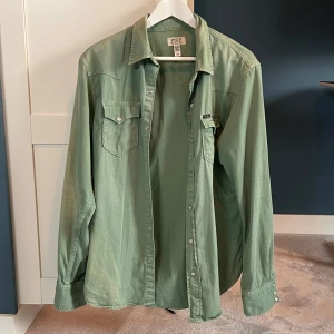 Vintage overshirt | Polo Ralph Lauren - Snygg grön skjorta från Polo Ralph Lauren i bomull. Klassisk westernstil med tryckknappar, två bröstfickor med lock och lång ärm. Skjortan har en avslappnad passform och diskret Polo-märke på ena fickan. Perfekt nu för hösten. Shortan är även solblekt en aning vilket gör den snyggare   